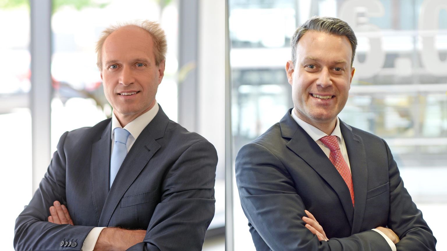 Two professionally dressed men are standing side by side, each with their arms crossed and smiling. The man on the left has blonde hair and a light blue tie, while the man on the right has dark hair and a red tie. The background features blurred glass and natural light.