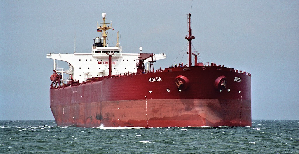 A large red cargo ship named "Molda" is sailing on the ocean. The vessel features a prominent bridge and is surrounded by water. The sky appears overcast, indicating a cloudy day. The ship has various structures and equipment visible on its deck.