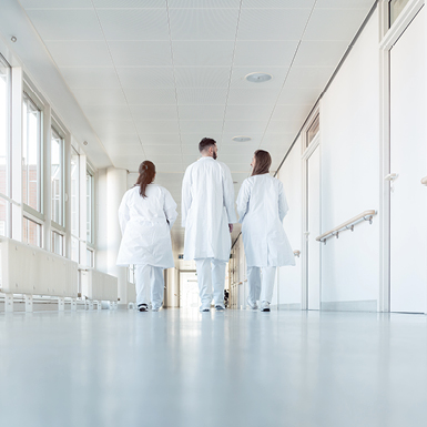 Drei Personen in weißen Laborkitteln gehen den Flur eines Krankenhauses entlang. Die Ansicht ist von hinten, mit großen Fenstern zu beiden Seiten. Die Atmosphäre wirkt hell und professionell.