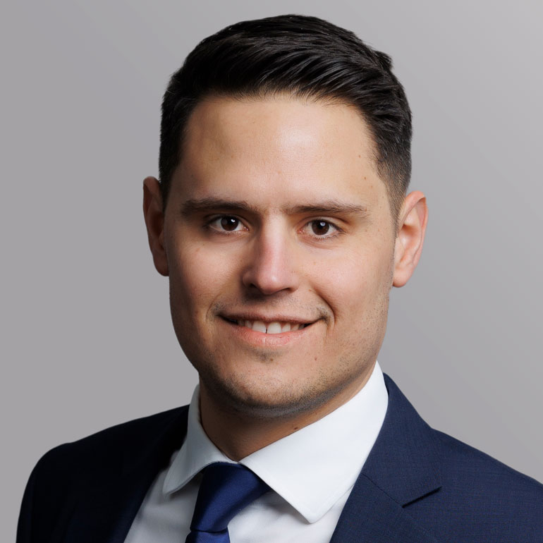 Porträt eines Mannes mit kurzem, glattem Haar, der in einem dunkelblauen Anzug und einer hellen Krawatte gekleidet ist. Er lächelt freundlich in die Kamera. Der Hintergrund ist grau und neutral gehalten.