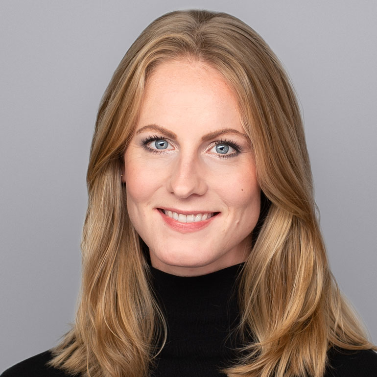 Das Bild zeigt eine junge Frau mit langen, blonden Haaren und einem freundlichen Lächeln. Sie trägt einen schwarzen Rollkragenpullover und blickt direkt in die Kamera. Der Hintergrund ist hellgrau, was ihr Gesicht betont.