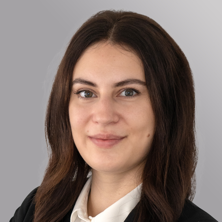 Einporträt einer jungen Frau mit langen, dunklen Haaren, die in einem schwarzen Blazer und einem weißen Oberteil gekleidet ist. Sie blickt direkt in die Kamera und hat einen freundlichen Ausdruck. Der Hintergrund ist grau und neutral gehalten.