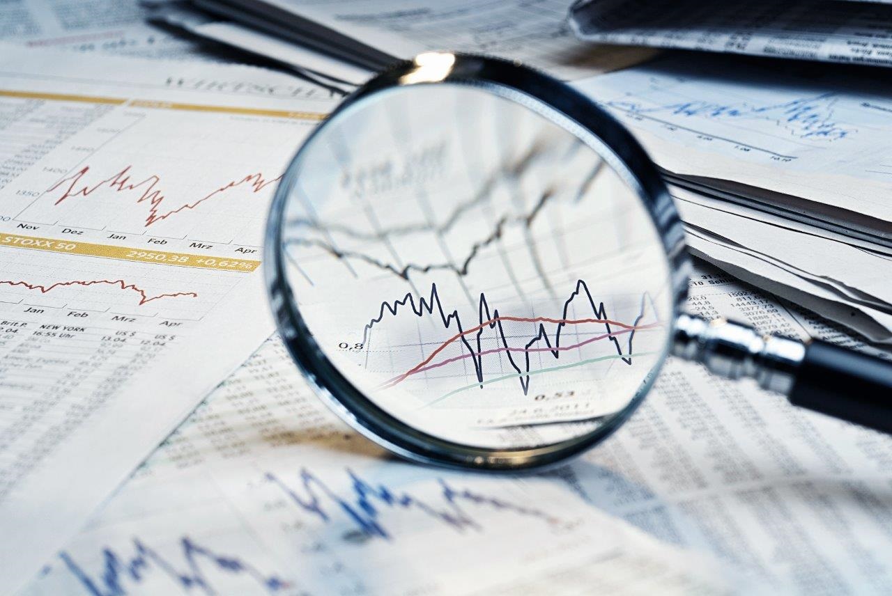 A magnifying glass focuses on financial charts and graphs displaying fluctuating trends in market data. Surrounding sheets of paper contain more detailed financial information, emphasizing analysis and interpretation of the data.