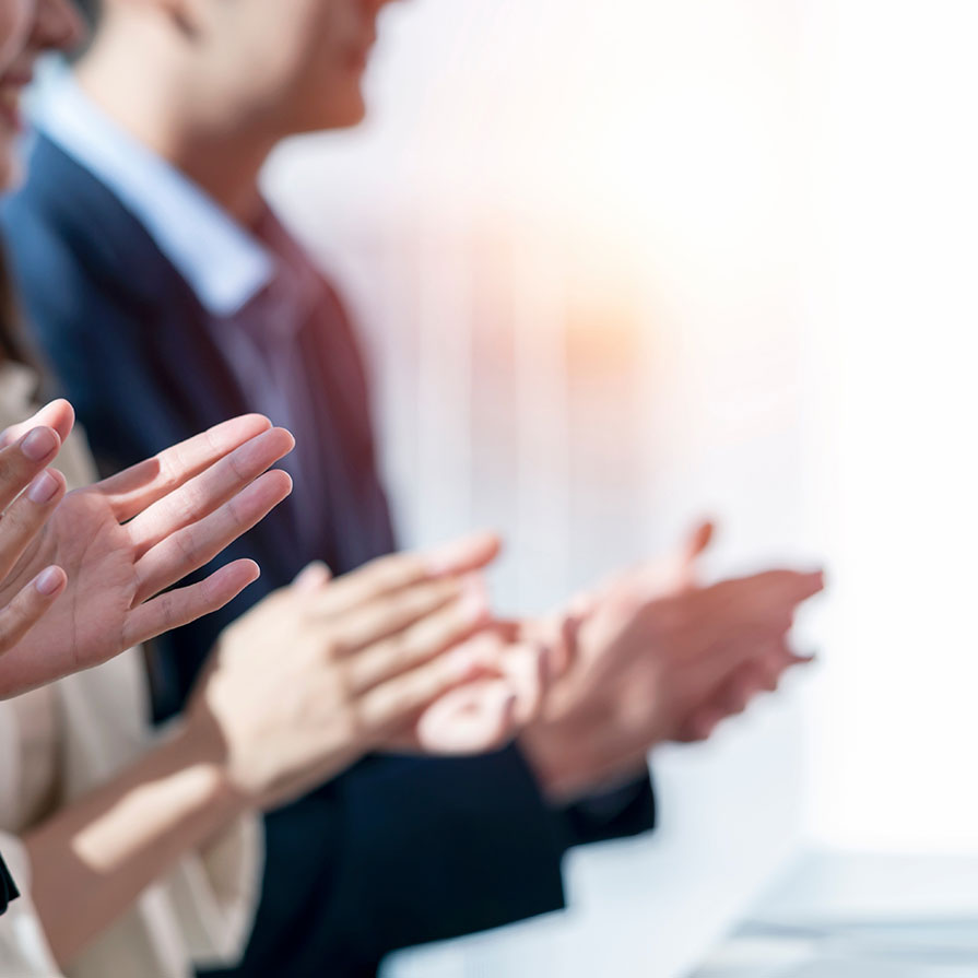 Einige Hände applaudieren in einer geschäftlichen Umgebung. Die Personen stehen nebeneinander, sichtbar sind die Hände und teilweise die Oberkörper. Die Szene vermittelt eine positive Stimmung, wahrscheinlich als Reaktion auf eine Präsentation oder einen Vortrag.
