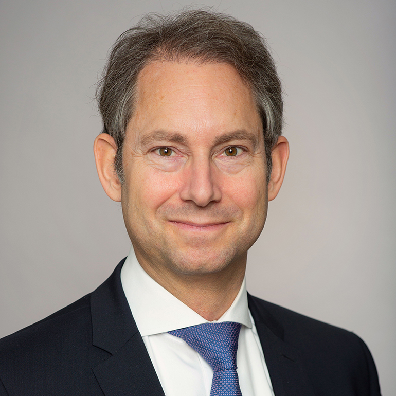 Ein Mann mit mittellangen, grauen Haaren lächelt freundlich in die Kamera. Er trägt einen dunklen Anzug, ein weißes Hemd und eine blauen Krawatte. Der Hintergrund ist hell und neutral.