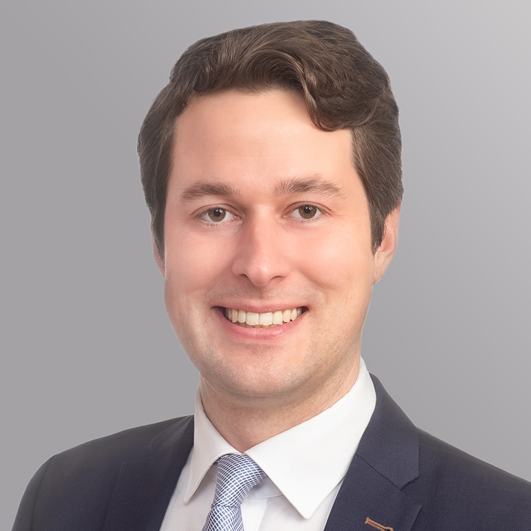 Ein junger Mann mit dunklen, lockigen Haaren lächelt freundlich. Er trägt einen dunkelblauen Anzug mit einem weißen Hemd und einer hellblauen Krawatte. Der Hintergrund ist grau.