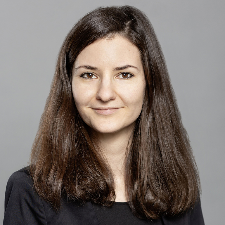 Porträt einer jungen Frau mit langen, braunen Haaren und einem freundlichen Lächeln. Sie trägt ein schwarzes Oberteil und hat einen neutralen Hintergrund. Die Aufnahme vermittelt einen professionellen und positiven Eindruck.