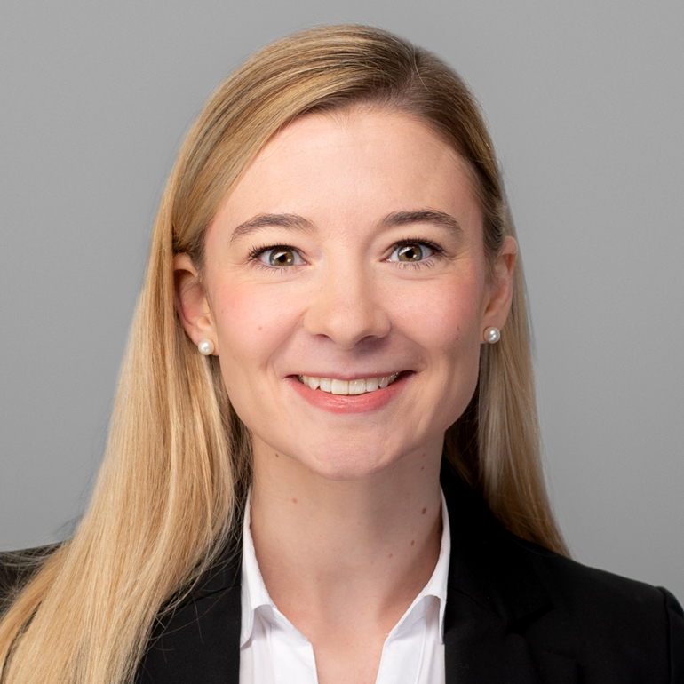 Ein Porträt einer Frau mit langen, blonden Haaren. Sie trägt ein schwarzes Jacket und eine weiße Bluse. Sie lächelt freundlich und blickt direkt in die Kamera, vor einem grauen Hintergrund.