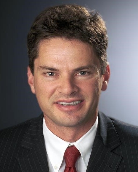 A smiling man with short, dark hair wearing a black pinstripe suit and a white shirt with a red tie. He is facing the camera against a neutral background, exuding a professional appearance.