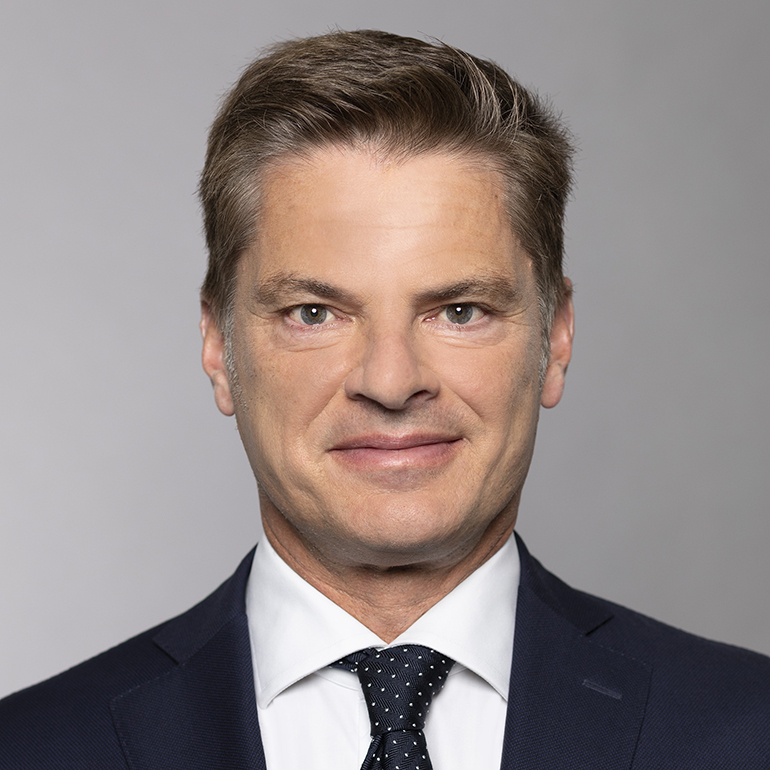 Ein Mann mit kurzen, dunkelblonden Haaren und einem leicht lächelnden Gesicht blickt direkt in die Kamera. Er trägt einen dunkelblauen Anzug und ein weißes Hemd mit einer gepunkteten Krawatte. Der Hintergrund ist neutral und hell.