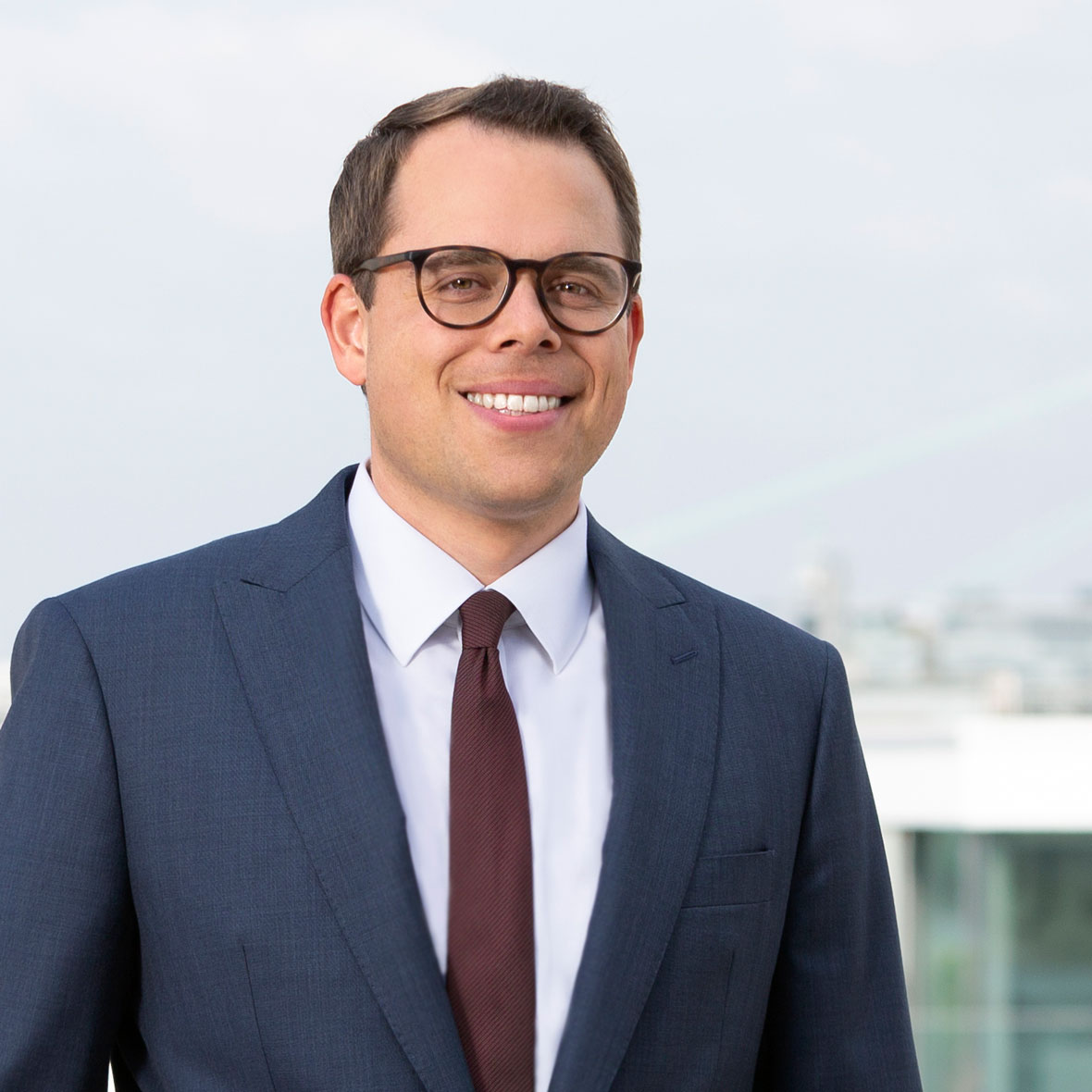 Ein gut gekleideter Mann mit Brille in einem Anzug steht lächelnd im Freien. Im Hintergrund sind unscharfe Gebäudestrukturen und ein heller Himmel sichtbar.