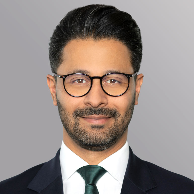 Ein junger Mann mit dunklem Haar und Bart trägt eine Brille und einen schwarzen Anzug mit einer weißen Hemd und einer grünen Krawatte. Er lächelt freundlich in die Kamera. Der Hintergrund ist grau und schlicht.