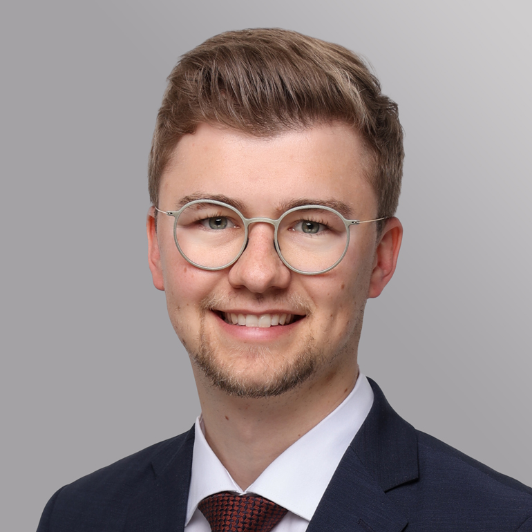 Ein junger Mann mit Brille lächelt freundlich in die Kamera. Er trägt einen dunklen Anzug und ein weißes Hemd mit einer roten Krawatte. Der Hintergrund ist in sanften Grautönen gehalten.