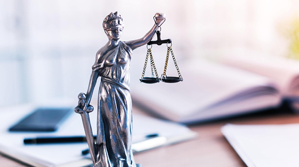 Eine silberne Statue der Justitia mit Augenbinde, die eine Waage in der einen Hand und ein Schwert in der anderen hält. Sie symbolisiert Gerechtigkeit und Rechtsprechung. Der Hintergrund zeigt verschwommene Notizen und Schreibutensilien.