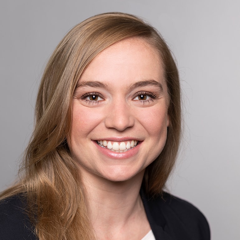 Ein Portrait einer lächelnden Frau mit langen, blonden Haaren und einem blauen Blazer vor einem neutralen Hintergrund. Sie hat einen freundlichen Gesichtsausdruck und wirkt selbstbewusst.