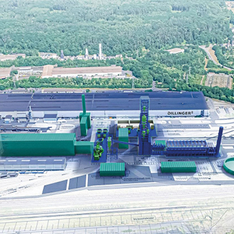 Luftaufnahme eines Industriekomplexes mit großen Gebäuden und Produktionsanlagen. Die Anlage, umgeben von Bäumen, besitzt mehrere grüne Container und hohe Türme. Der Name "Dillinger" ist auf einem der Gebäude sichtbar, was auf eine Stahlproduktionsstätte hinweist.