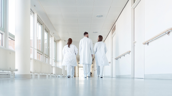 Drei Personen in weißen Laborkitteln gehen einen hellen Krankenhausflur entlang. Die Umgebung ist modern und gut beleuchtet, mit großen Fenstern, die Tageslicht hereinscheinen.