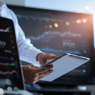 Eine Person in einem weißen Hemd hält ein Tablet in der Hand. Im Hintergrund ist ein Bildschirm mit Finanzgrafiken und Daten sichtbar, die sich auf das Trading oder die Analyse von Märkten beziehen.