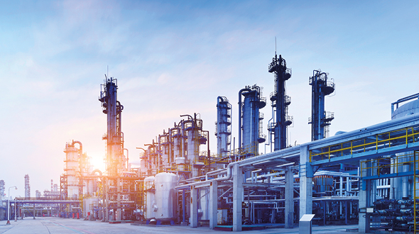 Industriegelände mit mehreren hohen Anlagen und Rohren, die in den Himmel ragen. Die Szene wird von warmen Sonnenstrahlen erleuchtet, die den frühen Morgen oder späten Nachmittag andeuten. Der Himmel ist klar und blau, was der industriellen Umgebung einen besonderen Kontrast verleiht.