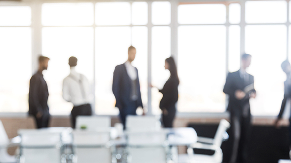 Eine Gruppe von sechs Menschen in formeller Kleidung steht in einem hellen Büro. Sie scheinen miteinander zu sprechen oder zu interagieren. Im Vordergrund sind Tisch und Stühle sichtbar, aber die Details sind unscharf.
