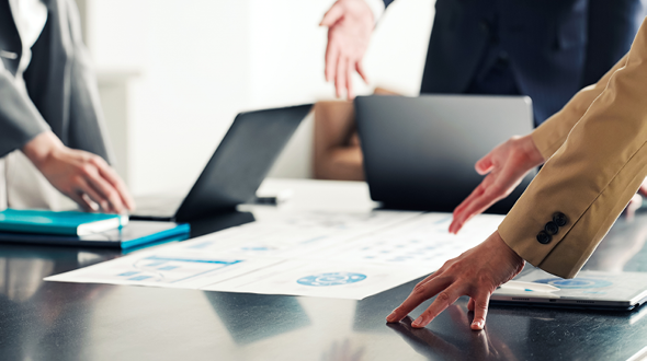 Das Bild zeigt einen Konferenzraum, in dem mehrere Personen um einen Tisch versammelt sind. Auf dem Tisch liegen Dokumente und Notizen, während einige Hände darauf zeigen oder gestikulieren. Im Hintergrund sind Laptops und eine bequeme Sitzgelegenheit sichtbar.