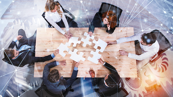 Eine Gruppe von sechs Personen sitzt um einen Tisch und hält ein großes, fehlendes Puzzlestück in der Mitte. Die Teilnehmer scheinen aktiv über die Lösung eines Problems nachzudenken und arbeiten zusammen, um die Puzzlestücke zu verbinden.