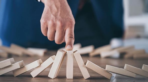 Ein Handfinger berührt einen dominosteinartigen Block, während mehrere ähnliche Blocks um ihn herum angeordnet sind. Die Szene symbolisiert Stabilität und das Verhindern eines Umkippens oder Zusammenbruchs.