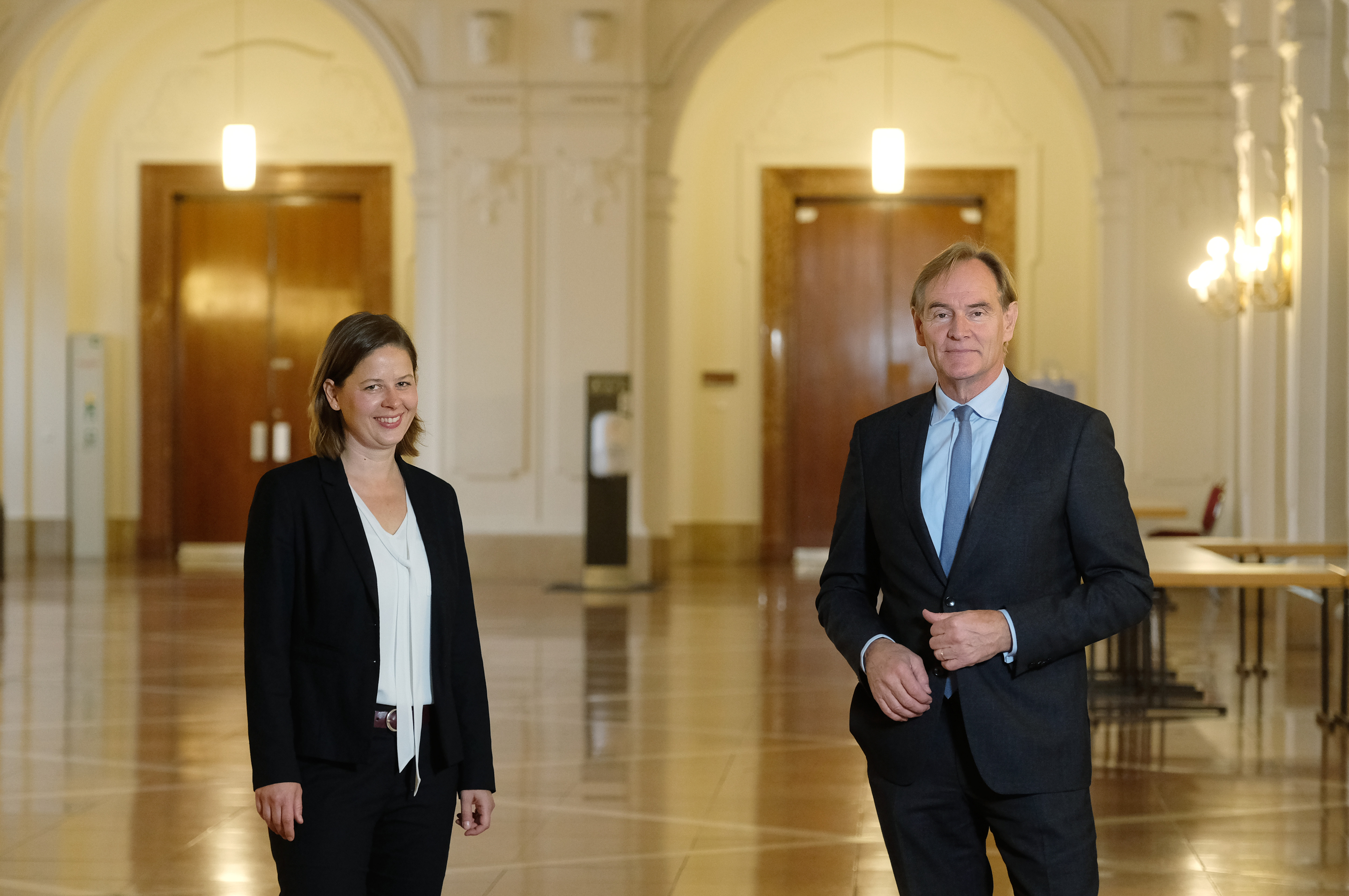 Two professionals stand in an elegant hall with arched doorways and polished floors. One is a woman in a black suit, smiling, and the other is a man in a dark suit with a light blue shirt, looking towards the camera.