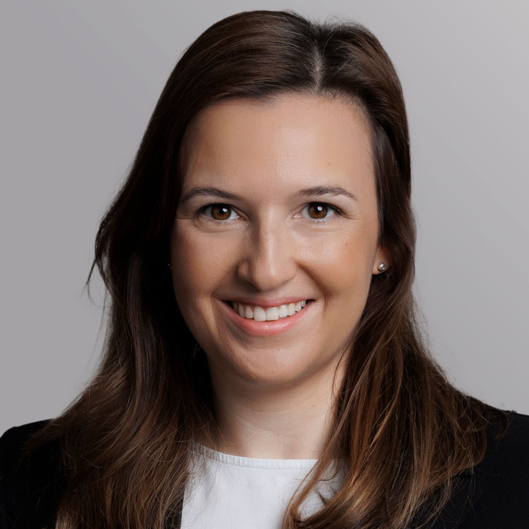 Porträt einer lächelnden Frau mit langen, bräunlichen Haaren. Sie trägt ein schwarzes Blazer-Oberteil und einen weißen Unterbau. Der Hintergrund ist grau. Ihr Ausdruck vermittelt Freundlichkeit und Offenheit.