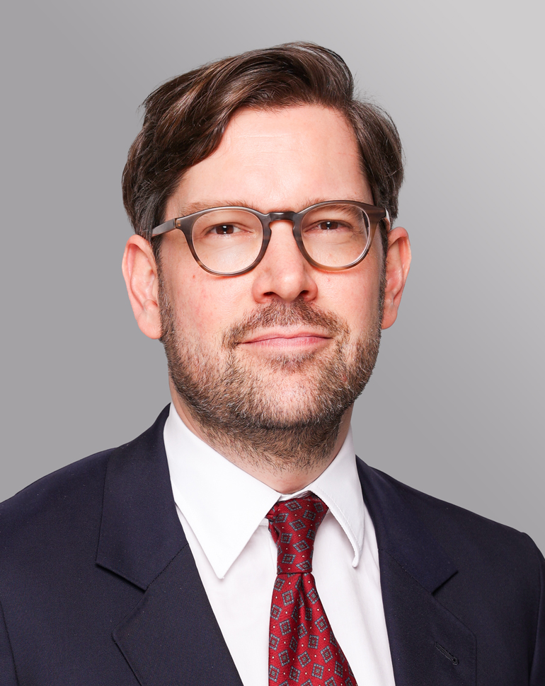 Portrait eines Mannes mit Brille, der einen Anzug und eine Krawatte trägt. Er schaut direkt in die Kamera und wirkt selbstbewusst. Der Hintergrund ist einfarbig und neutral.