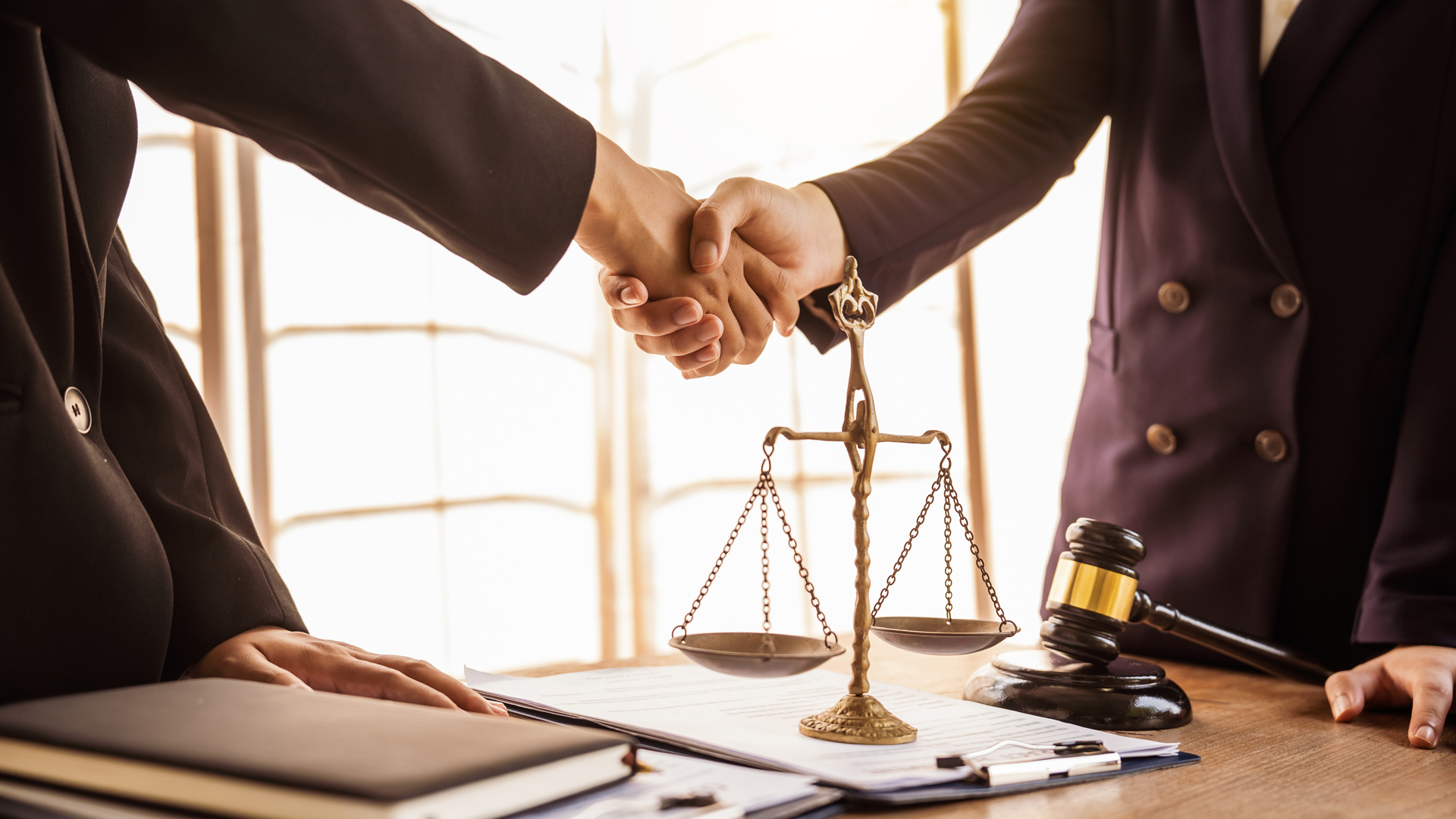 Zwei Personen schütteln sich die Hände über einem Tisch, auf dem eine Waage und ein Richterhammer liegen, was auf eine rechtliche Einigung oder Zusammenarbeit hinweist. Die Szene strahlt Professionalität und den Kontext eines rechtlichen Umfelds aus.