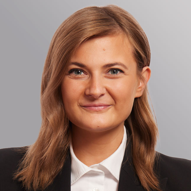 Ein Porträt einer jungen Frau mit braunen Haaren und einem freundlichen Lächeln. Sie trägt ein schwarzes Jacket und ein weißes Hemd. Der Hintergrund ist in sanften Grautönen gehalten.