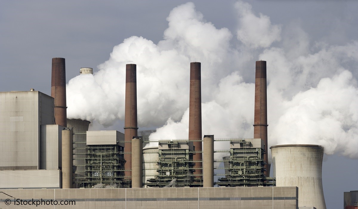 A large industrial facility features several tall smokestacks releasing white vapor into the air. The structure appears modern, with various machinery visible, suggesting it is a power plant or factory. The scene indicates an active operation contributing to environmental concerns.