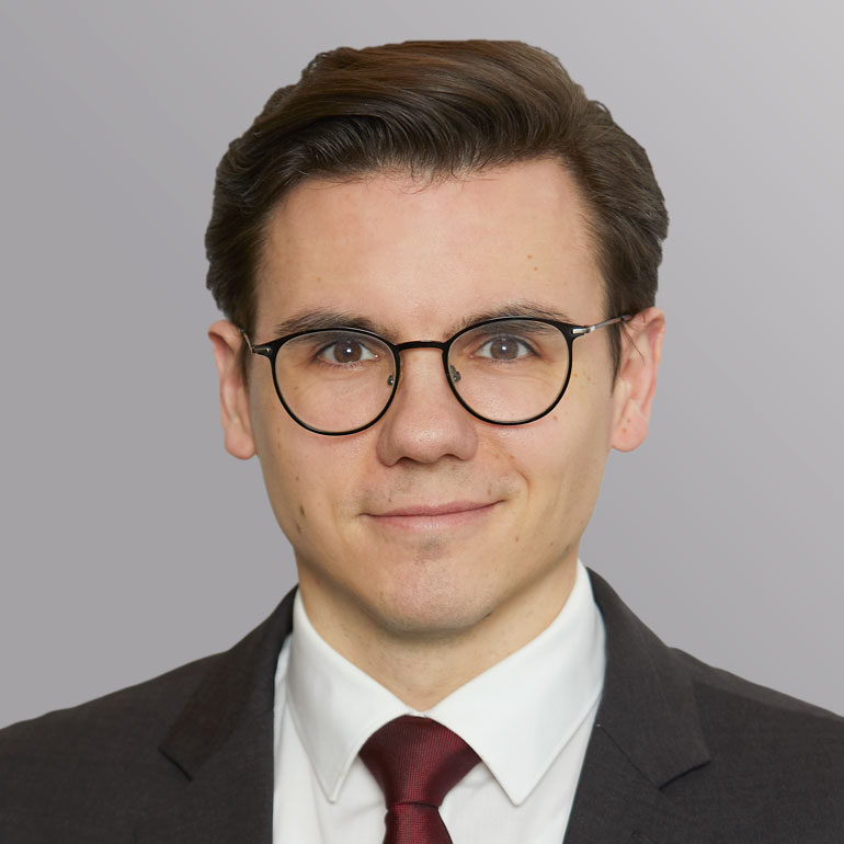 Porträt eines jungen Mannes mit Brille, der ein schwarzes Sakko und ein weißes Hemd mit einer roten Krawatte trägt. Er lächelt freundlich vor einem grauen Hintergrund.