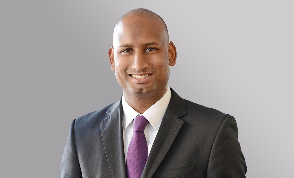 Ein lächelnder Mann in einem schwarzen Anzug mit einem weißen Hemd und einer lila Krawatte. Der Hintergrund ist grau. Die Person vermittelt einen freundlichen und professionellen Eindruck.