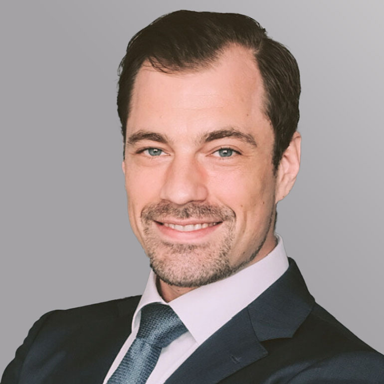 Porträt eines lächelnden Mannes in einem Anzug. Er hat kurze, dunkle Haare und trägt ein helles Hemd mit einer Krawatte. Der Hintergrund ist grau. Der Mann wirkt freundlich und selbstbewusst.