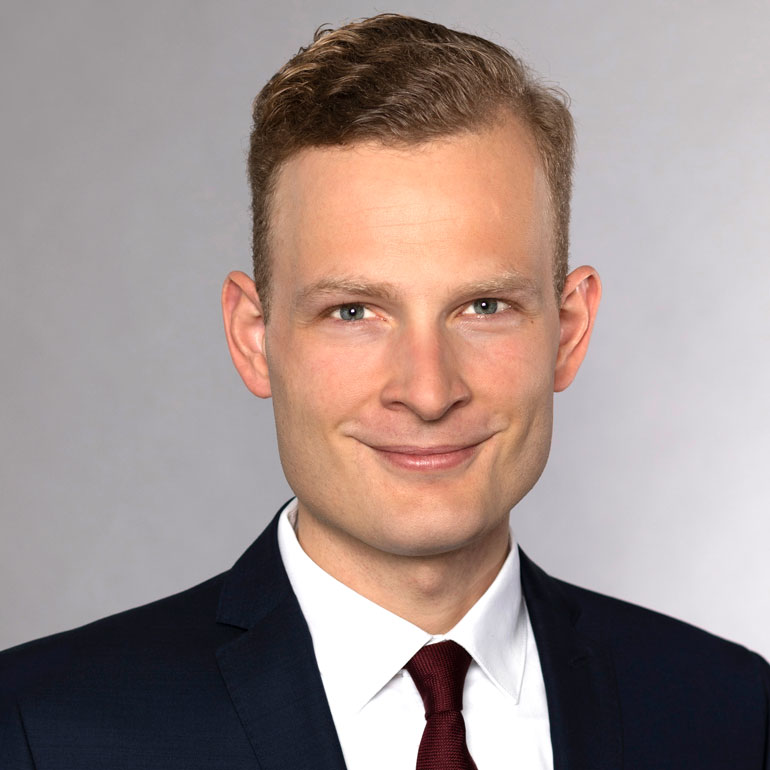 Porträt eines Mannes mit kurzen, blonden Haaren und einem lächelnden Gesichtsausdruck. Er trägt einen dunklen Anzug mit einer weißen Hemd und einer roten Krawatte. Der Hintergrund ist hell und neutral.