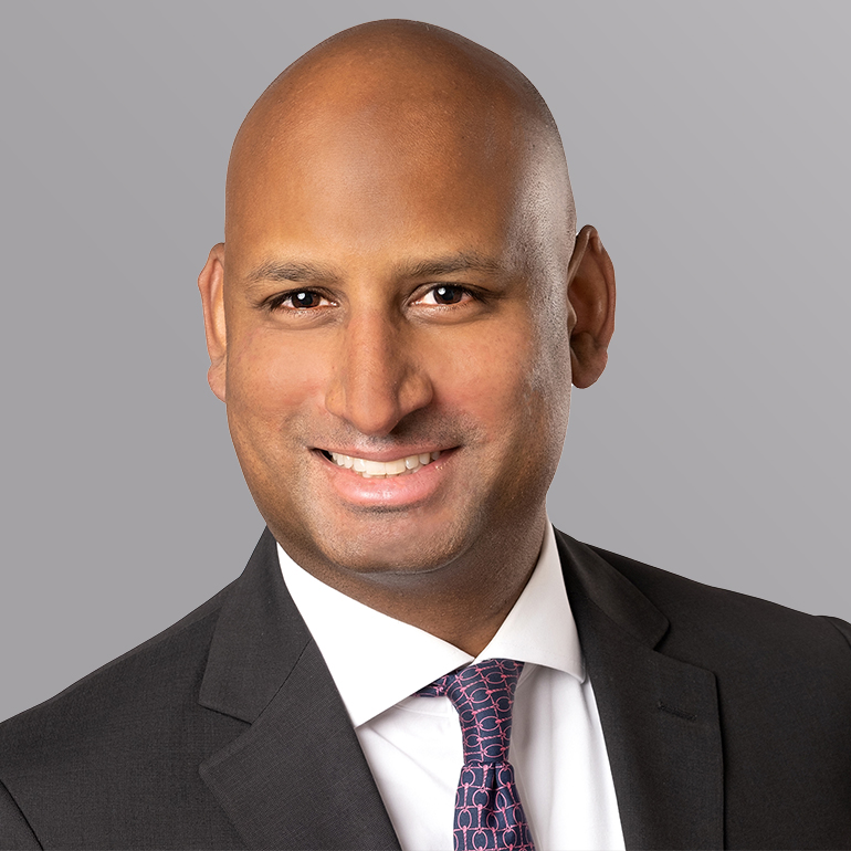 Ein lächelnder Mann mit kahlem Kopf trägt einen Anzug und eine Krawatte mit einem geometrischen Muster. Der Hintergrund ist grau. Sein Ausdruck vermittelt Freundlichkeit und Professionalität.