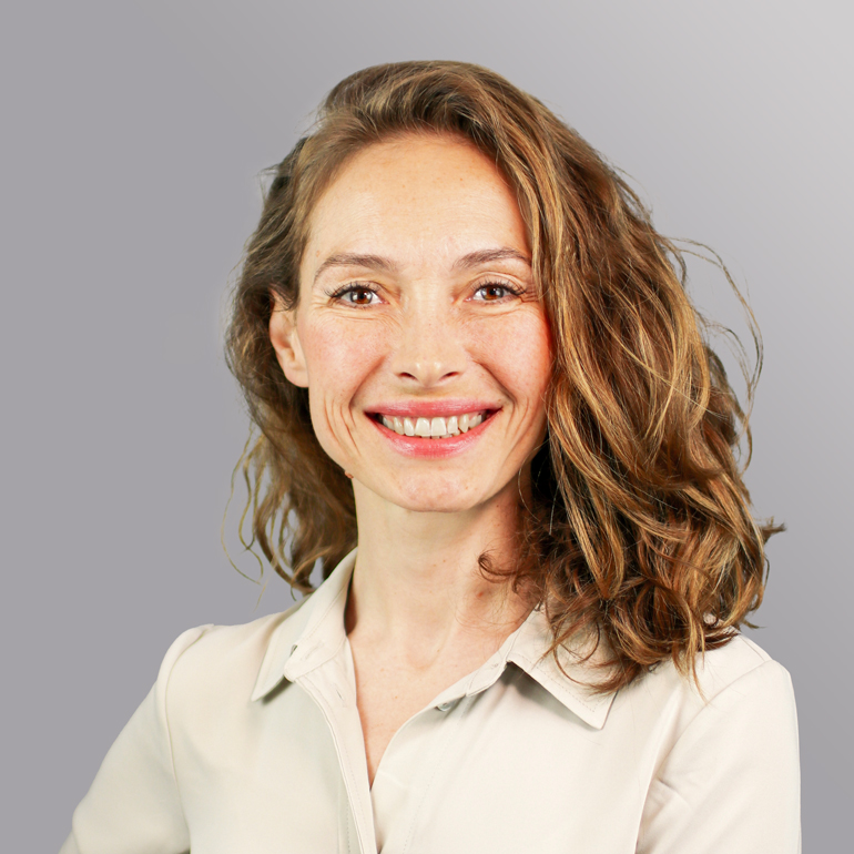 Porträt einer lächelnden Frau mit lockigem, brünettem Haar. Sie trägt ein helles Hemd und hat einen freundlichen Ausdruck. Der Hintergrund ist grau und leicht verschwommen, was den Fokus auf ihr Gesicht lenkt.