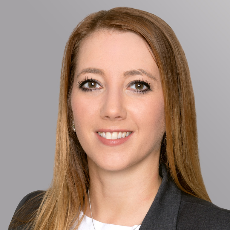 Ein Porträt einer Frau mit langen, braunen Haaren und einem freundlichen Lächeln. Sie trägt einen dunklen Blazer und steht vor einem grauen Hintergrund. Ihre Augen sind gut betont, und sie blickt direkt in die Kamera.