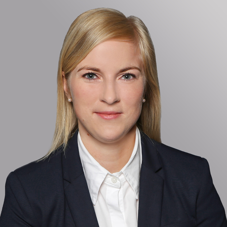 Ein Porträt einer Frau mit blonden, schulterlangen Haaren, die in einem dunklen Anzug und hellen Hemd gekleidet ist. Sie lächelt leicht und schaut direkt in die Kamera. Der Hintergrund ist grau und neutral gehalten.