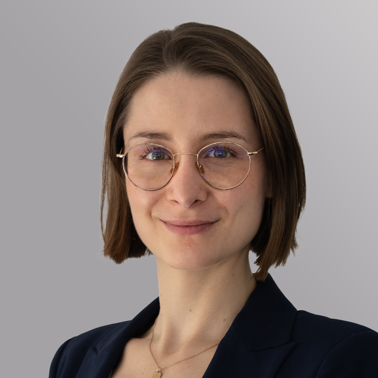 Porträt einer Frau mit kurzen, braunen Haaren und einer leichten Welle. Sie trägt eine dunkelblaue Jacke und runde, goldene Brille. Ihr Gesicht zeigt ein freundliches Lächeln, im Hintergrund ist ein einfarbiger, heller Farbverlauf zu sehen.