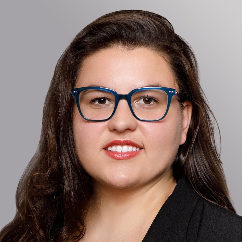 Porträt einer Frau mit langen, lockigen Haaren und blauen Brillen. Sie hat einen freundlichen Gesichtsausdruck, trägt einen schwarzen Blazer und hat einen natürlichen Lippenstift aufgetragen. Der Hintergrund ist grau und neutral gehalten.