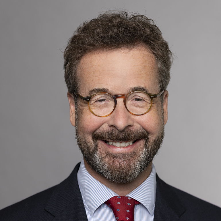 Porträt eines Mannes mit Brille, dunklem, lockigem Haar und Bart. Er trägt einen dunkelblauen Anzug mit einem hellblauen Hemd und einer rotem Krawatte. Der Mann lächelt freundlich vor einem neutralen, grauen Hintergrund.