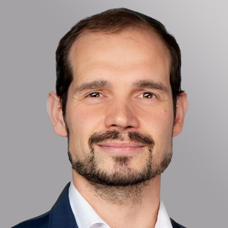 Ein lächelnder Mann mit kurzem, braunem Haar und Bart trägt ein dunkelblaues Jackett über einem weißen Hemd. Der Hintergrund ist einfarbig grau. Sein Gesichtsausdruck ist freundlich und zuversichtlich.