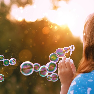 Ein Kind mit langen Haaren bläst Seifenblasen in den Sonnenuntergang. Die Blasen schweben durch die Luft und reflektieren das Licht. Der Hintergrund zeigt eine grüne Umgebung, die eine fröhliche, spielerische Atmosphäre vermittelt.
