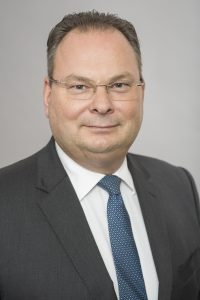 Ein Mann in Anzug mit Krawatte und Brille lächelt in die Kamera. Der Hintergrund ist neutral und hell.