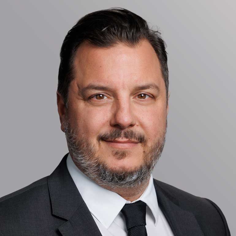 Ein Mann mit dunklen Haaren und einem gepflegten Bart lächelt in die Kamera. Er trägt einen Anzug mit Krawatte und ein Hemd. Der Hintergrund ist in sanften Grautönen gehalten.