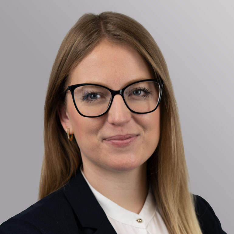 Porträt einer Frau mit langen, blonden Haaren und einer Bluse. Sie trägt große, schwarze Brille und lächelt leicht. Der Hintergrund ist grau. Das Bild vermittelt einen professionellen Eindruck.