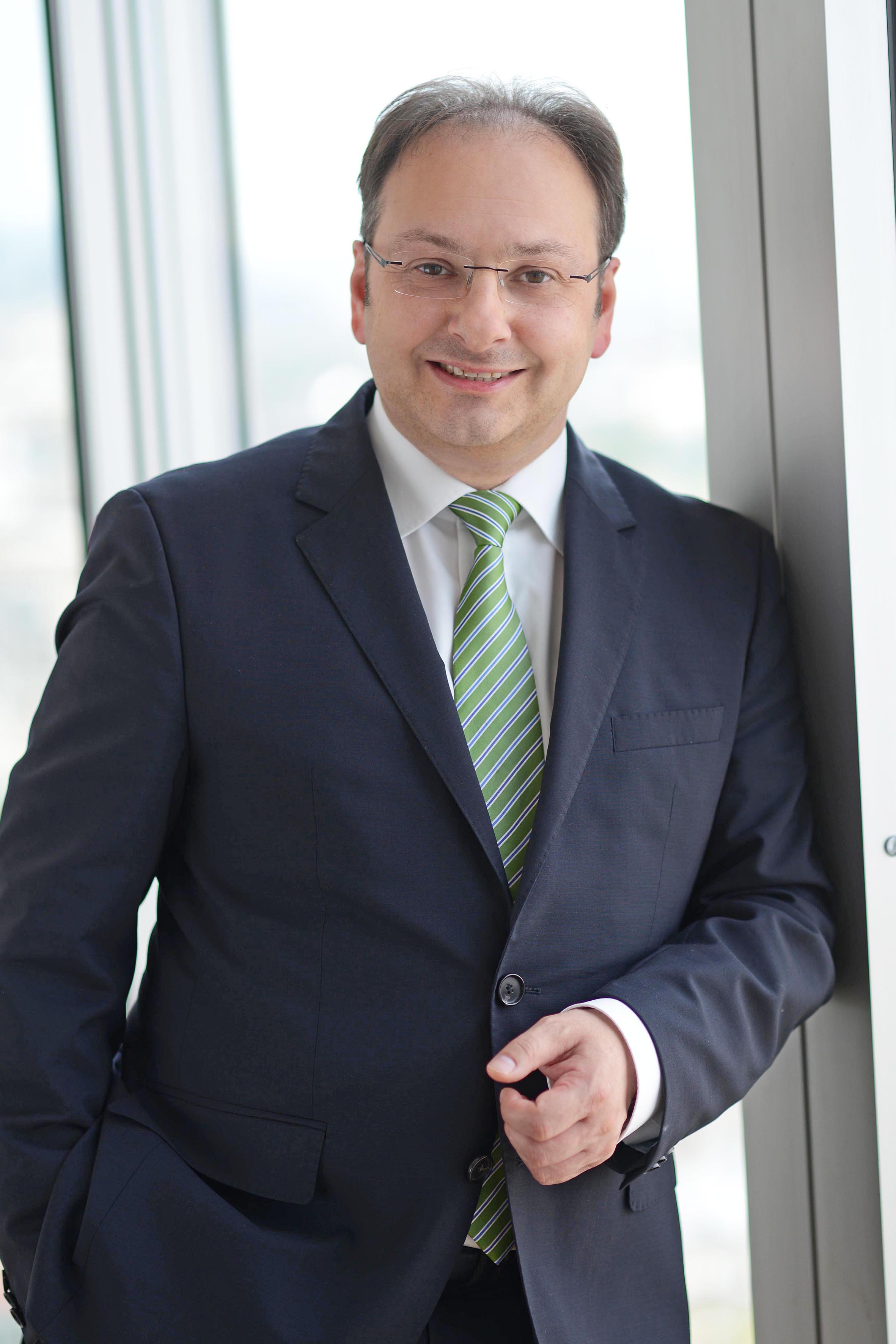 Ein Mann in einem dunkelblauen Anzug steht an einem Fenster. Er lächelt und trägt ein weißes Hemd mit einem grün gestreiften Krawatte. Seine Hände sind entspannt, eine Hand lehnt an der Wand. Der Hintergrund zeigt unscharfe Gebäude und Himmel.